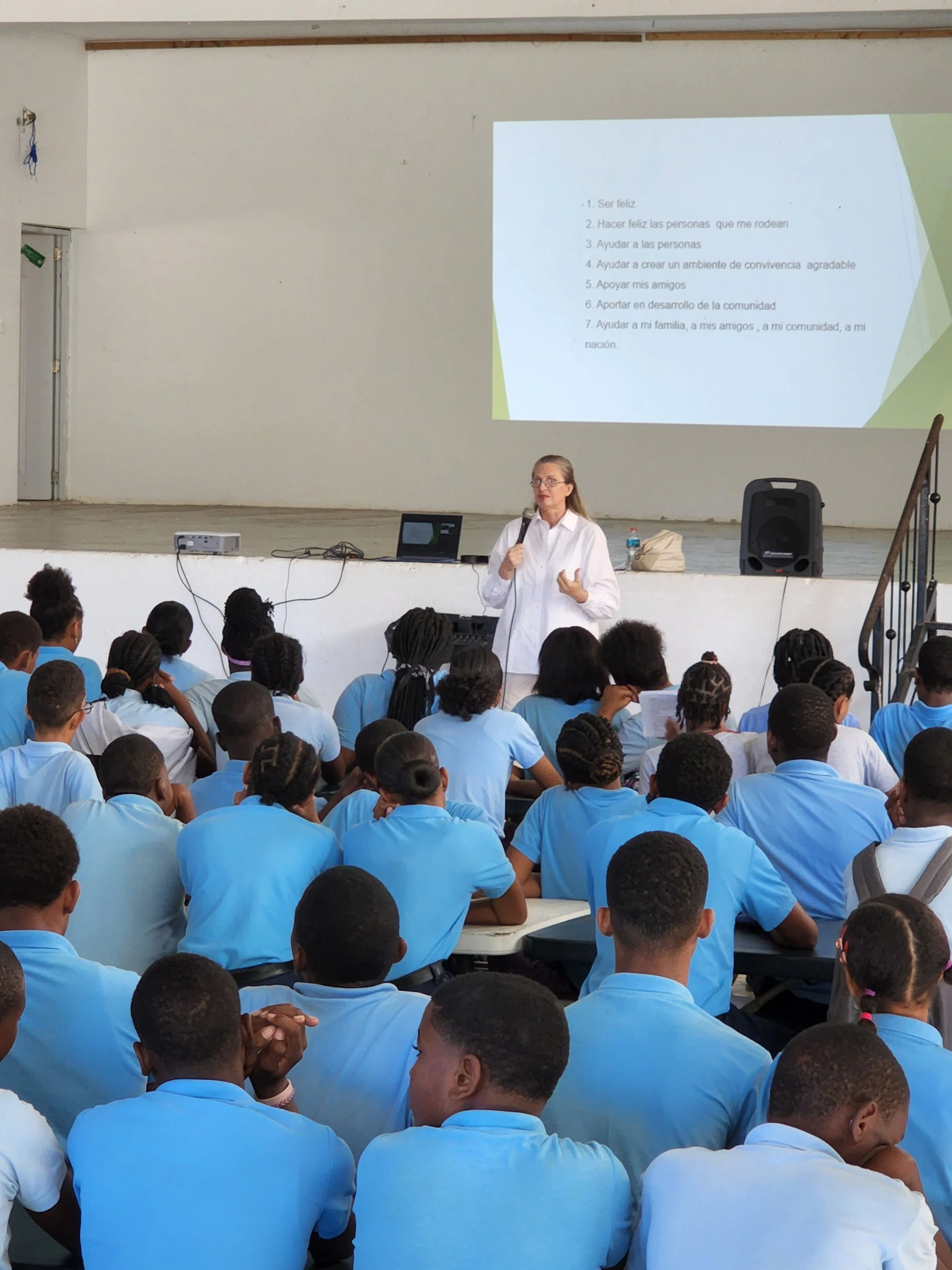 A woman in a white shirt giving a presentation to a group of students wearing blue shirts, seated on benches, in a classroom or auditorium setting. A large screen behind her displays text in Spanish.