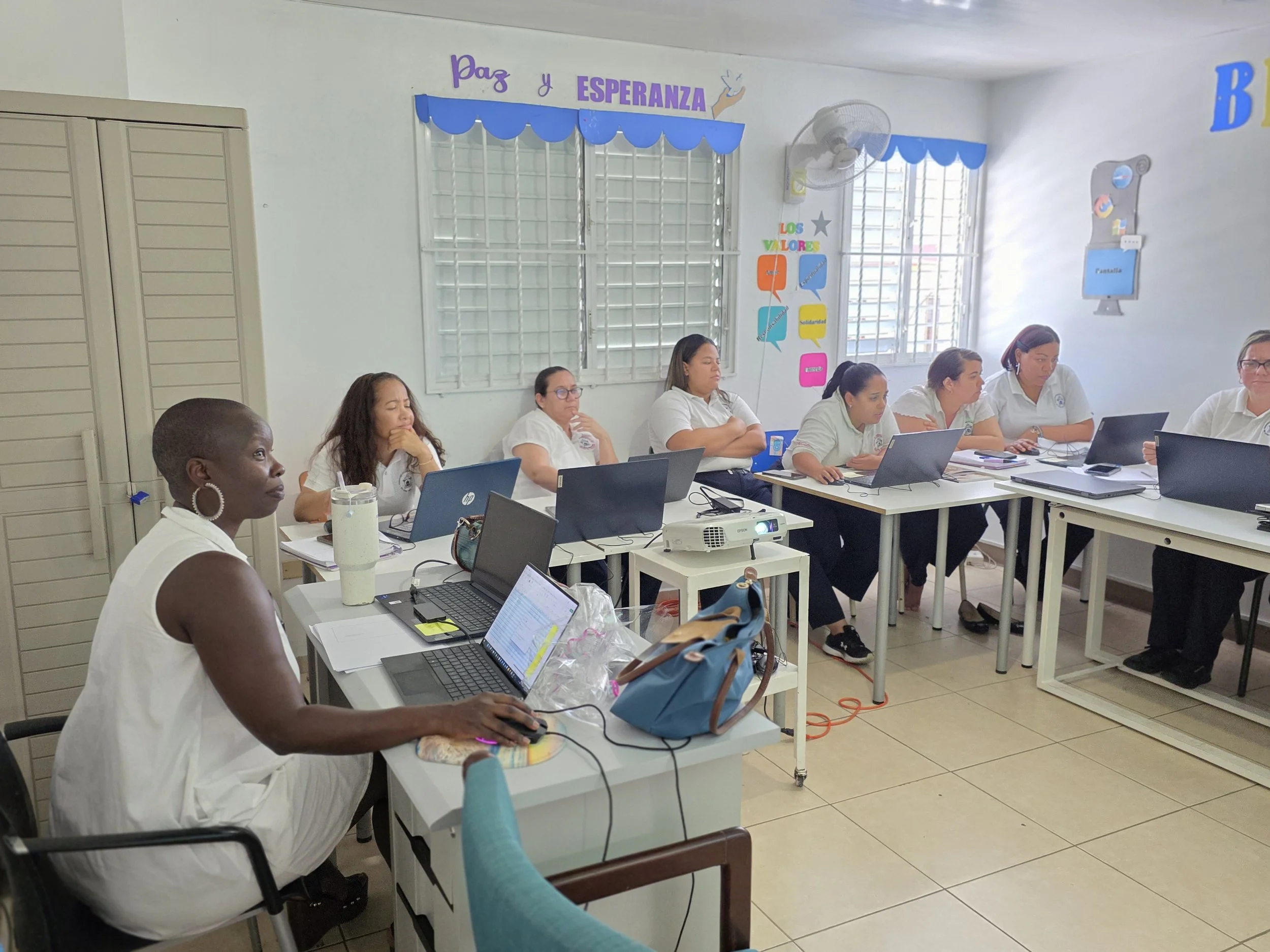 A group of women attending a meeting or workshop in a classroom. They are sitting at tables with laptops and notebooks. The room has educational posters on the walls with words in Spanish, and a large window with security bars.