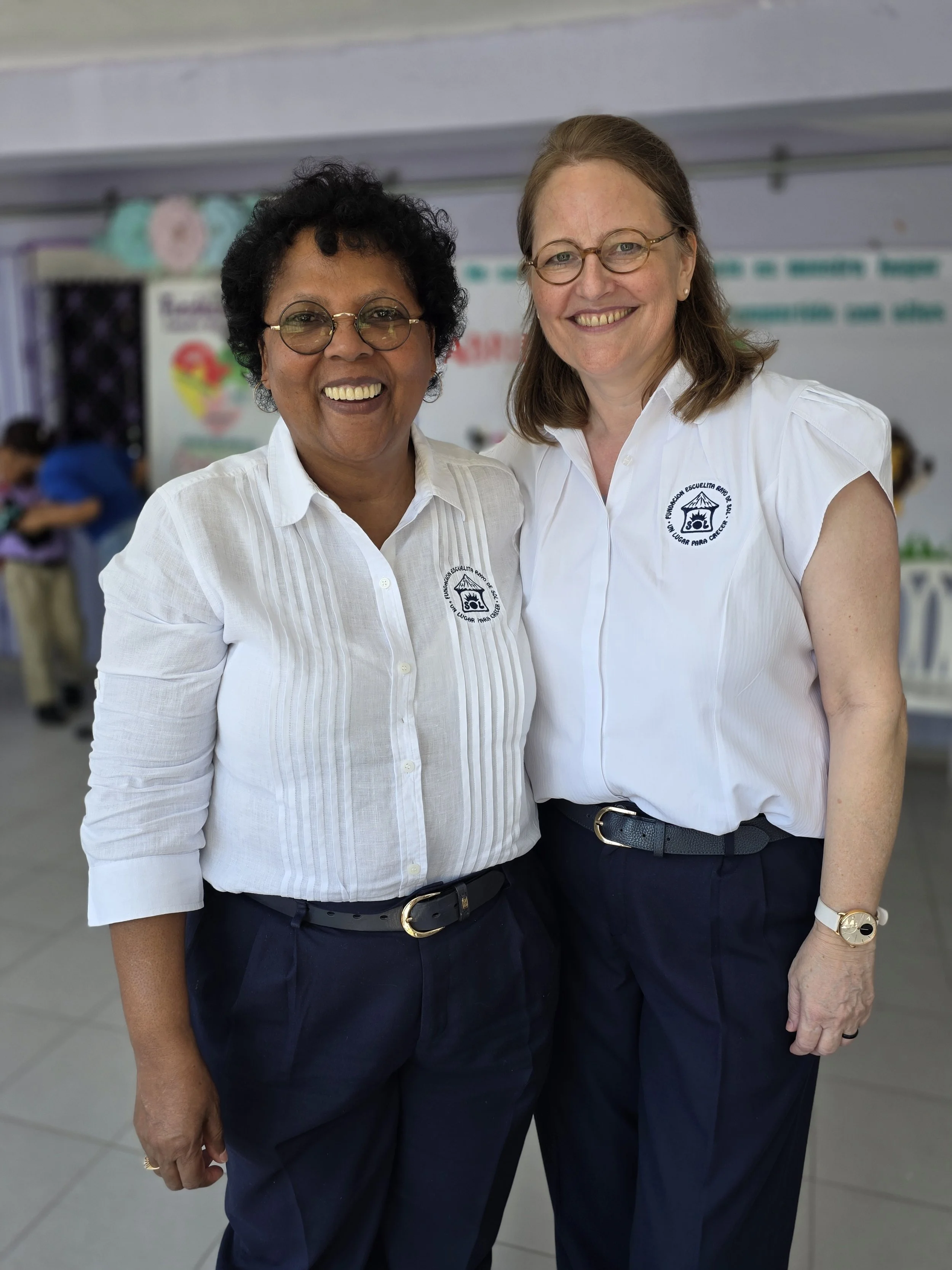 Two women smiling and standing close together, wearing white shirts with a school logo, dark pants, in a classroom or event setting.