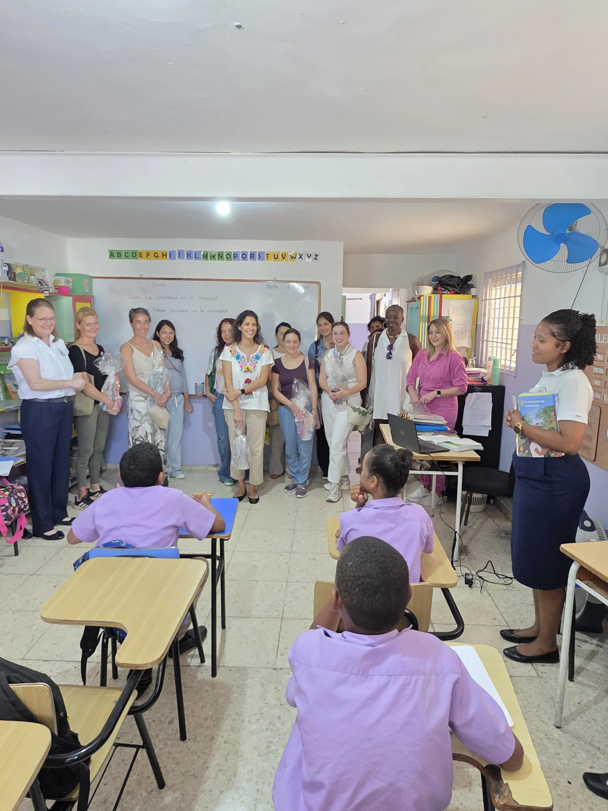 A classroom with students and teachers, some holding gift bags. Students are seated at desks, facing forward, while teachers stand at the front near a whiteboard and colorful alphabet border. The room has a blue fan on the wall and a window.