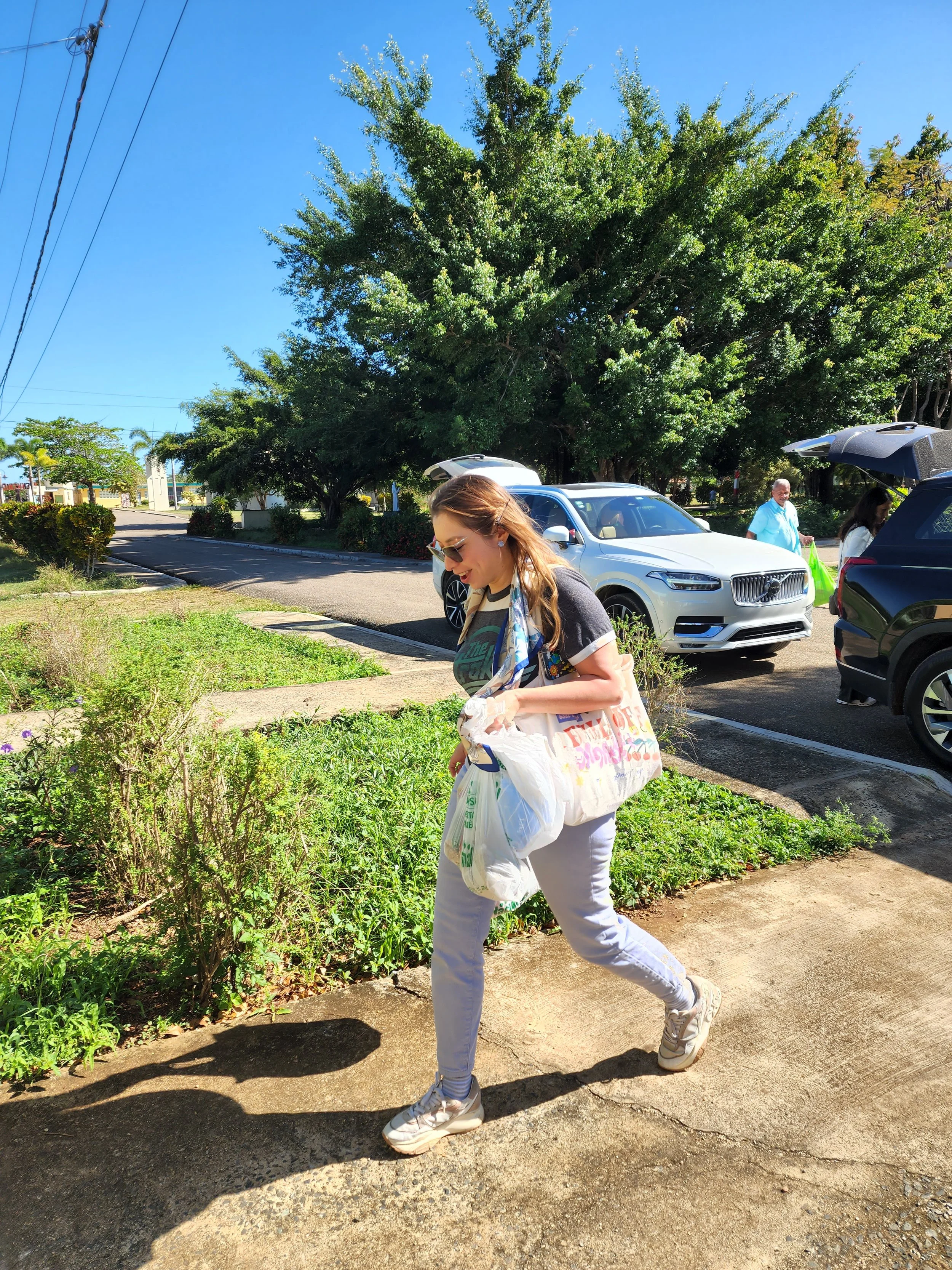 A woman with sunglasses walking on sidewalk, carrying shopping bags on her shoulder, enjoying a sunny day with a large green tree and parked cars in the background.