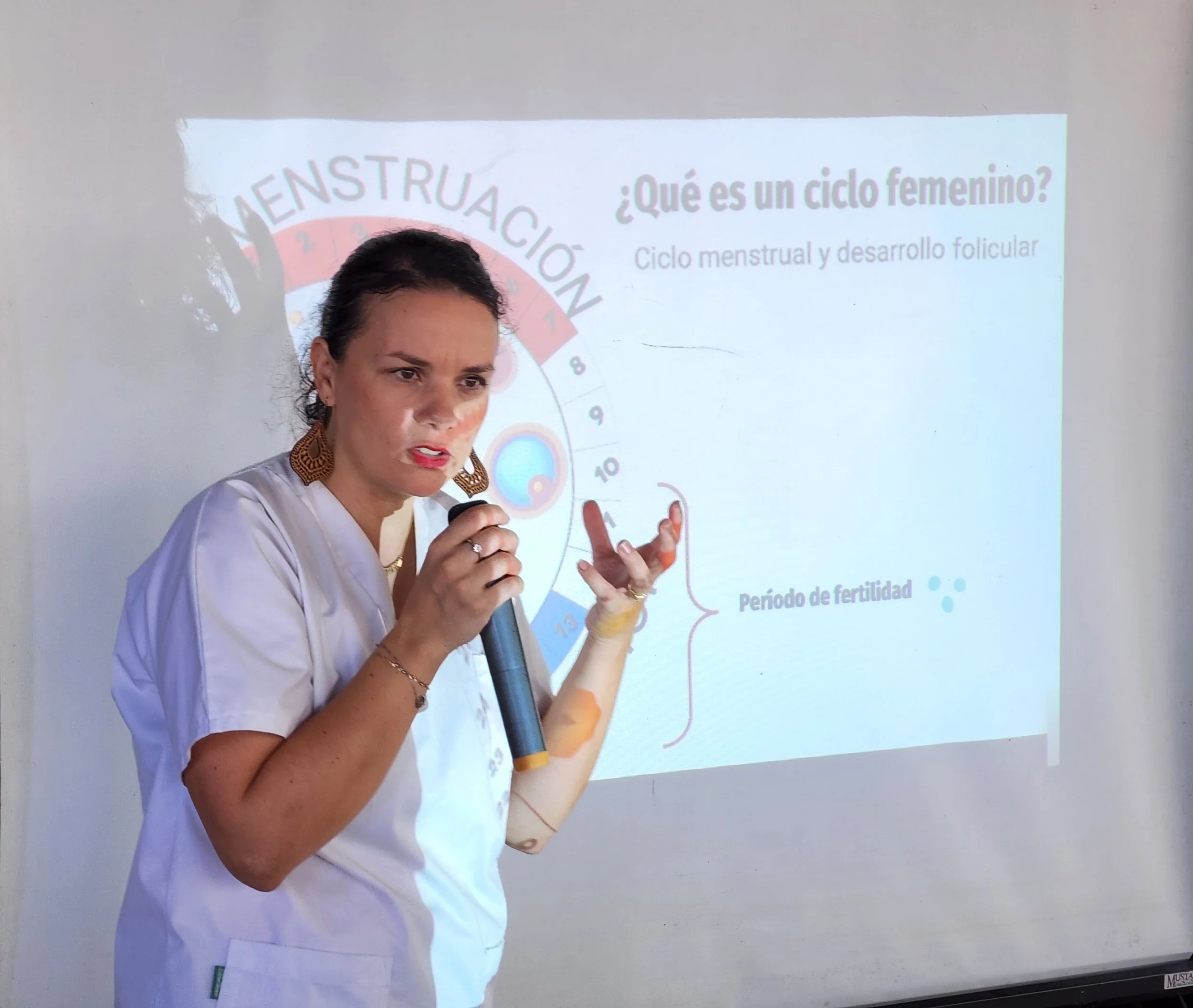 A woman is giving a presentation in front of a projected slide about the menstrual cycle, holding a microphone and gesturing with her hand. She is wearing a white uniform and earrings.