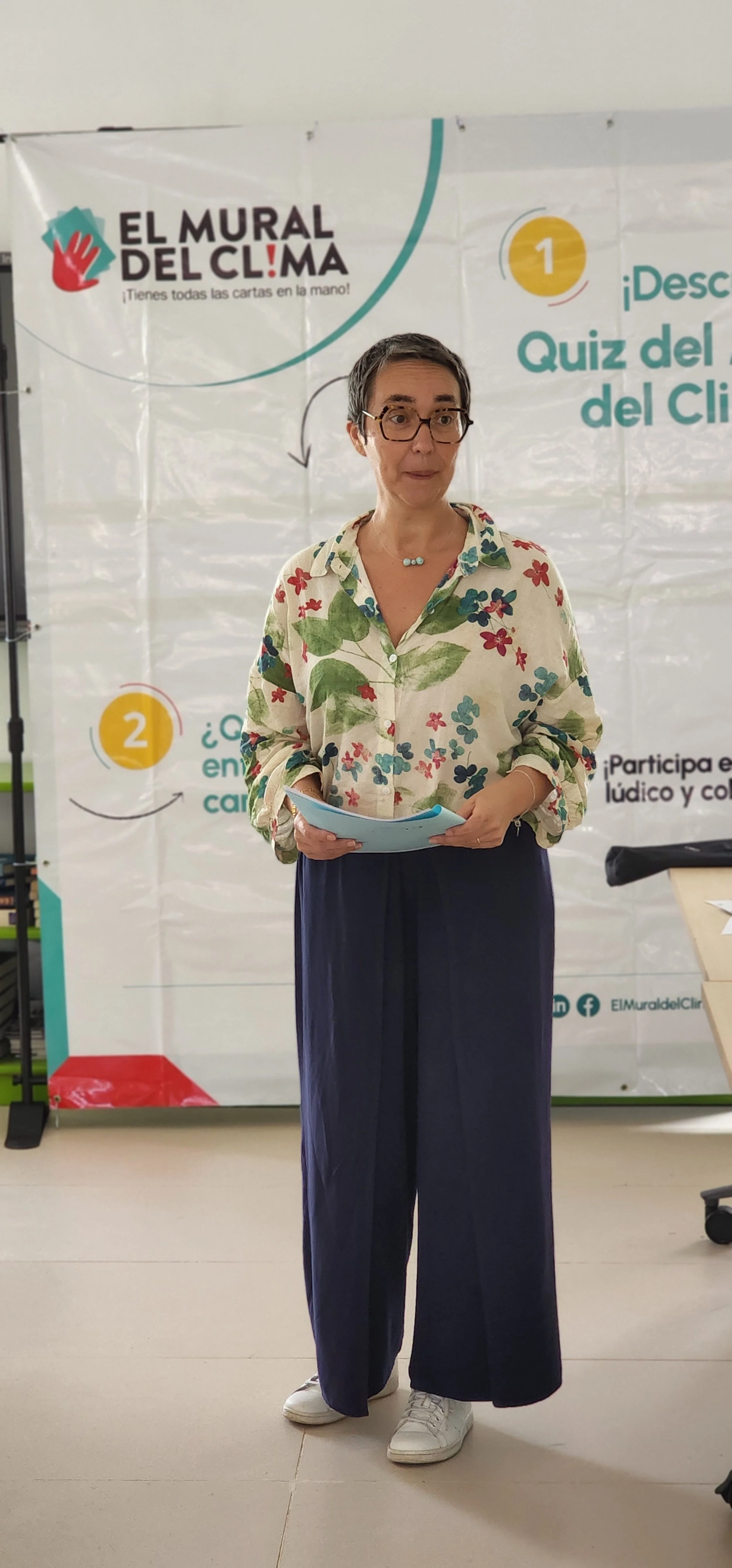 A woman with short gray hair, glasses, and a floral shirt standing in front of a white banner with colorful text and graphics, holding papers in her hands.
