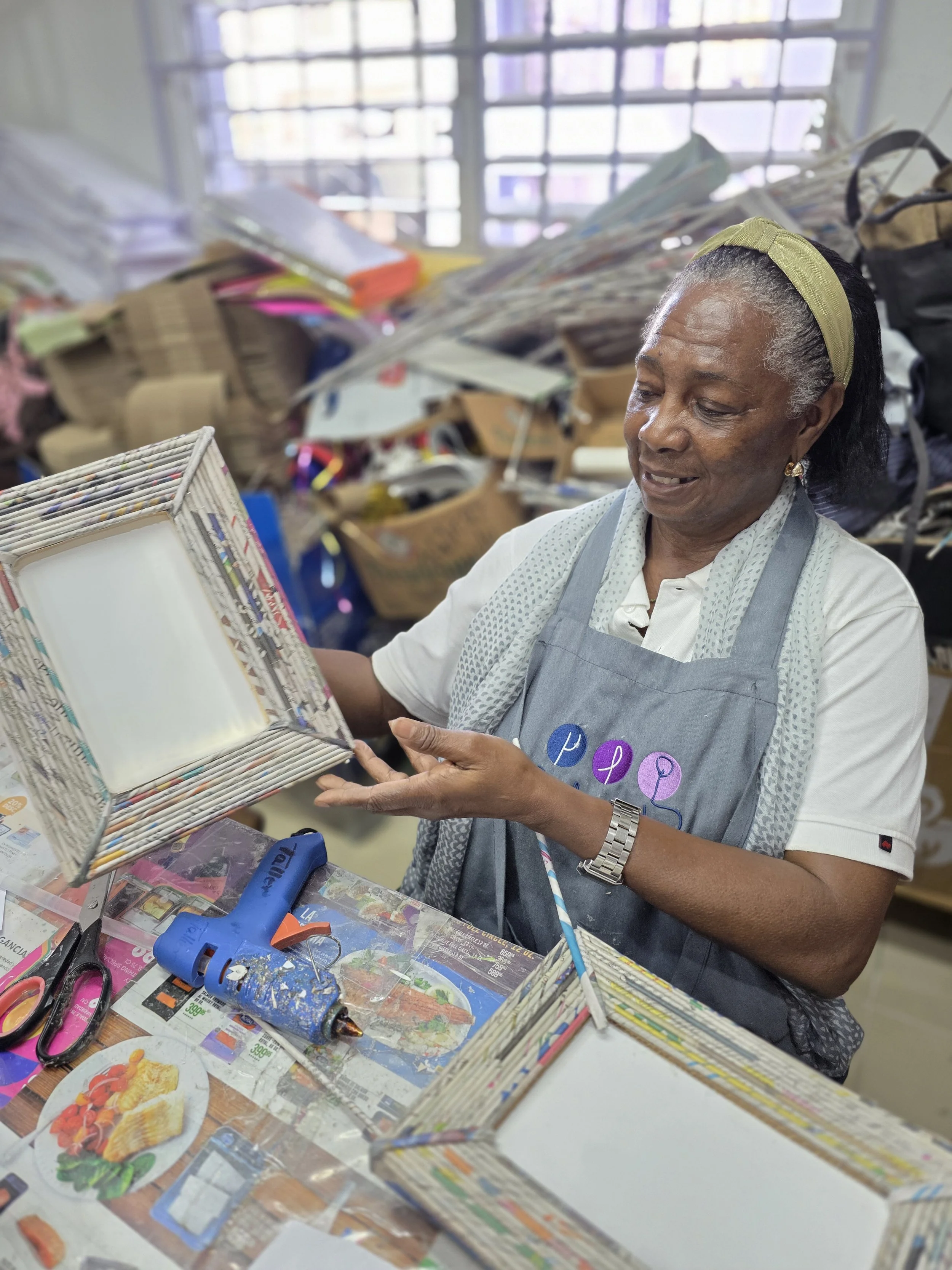 An elderly woman wearing a gray apron over a white shirt and a blue patterned scarf, working at a cluttered table covered with craft supplies, glue, scissors, and newspapers, holding a square frame made from rolled newspapers.