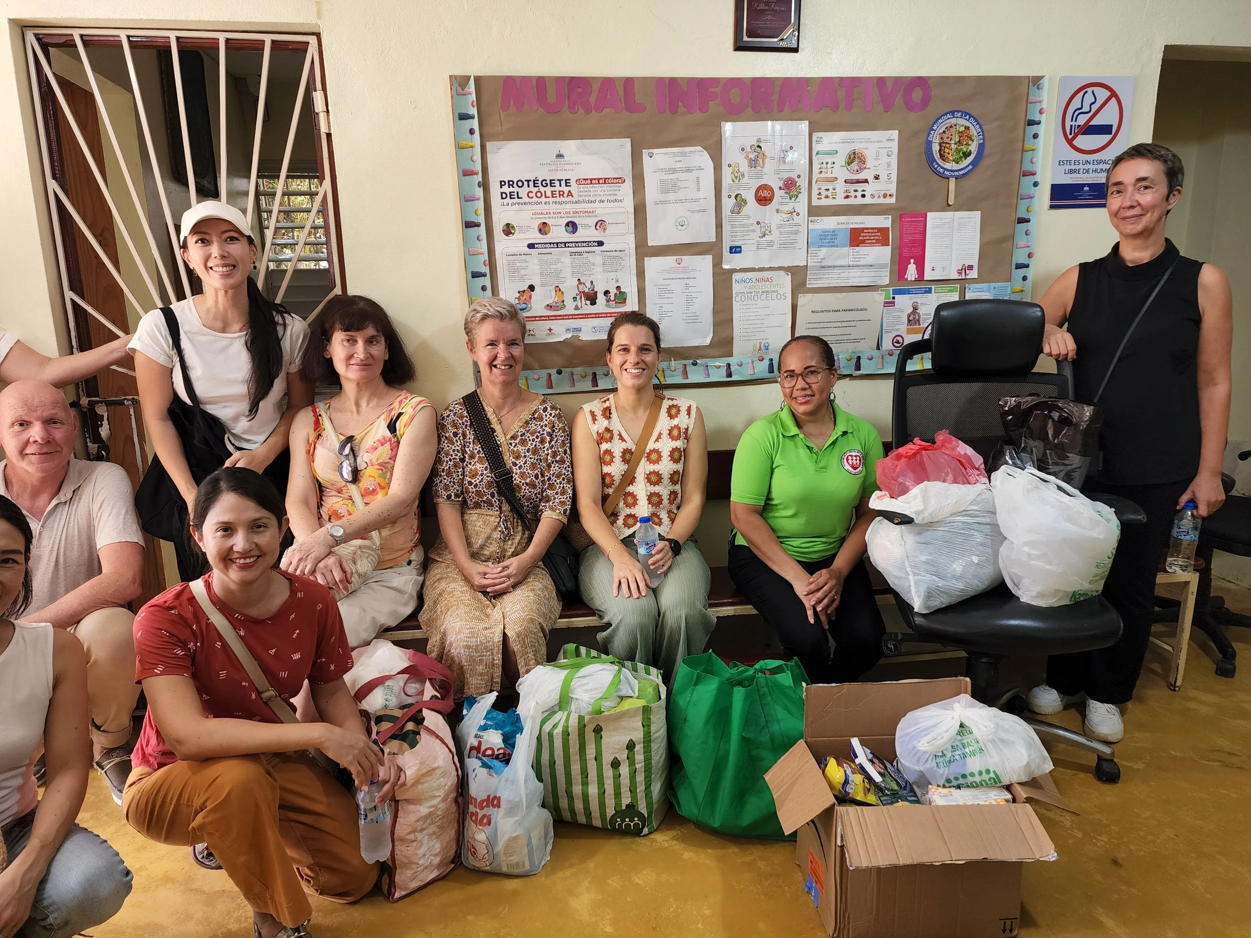 Group of ten people, mostly women, gathered in a room with a bulletin board that reads "MURAL INFORMATIVO" behind them. They are sitting and standing around bags and boxes filled with supplies or donations.