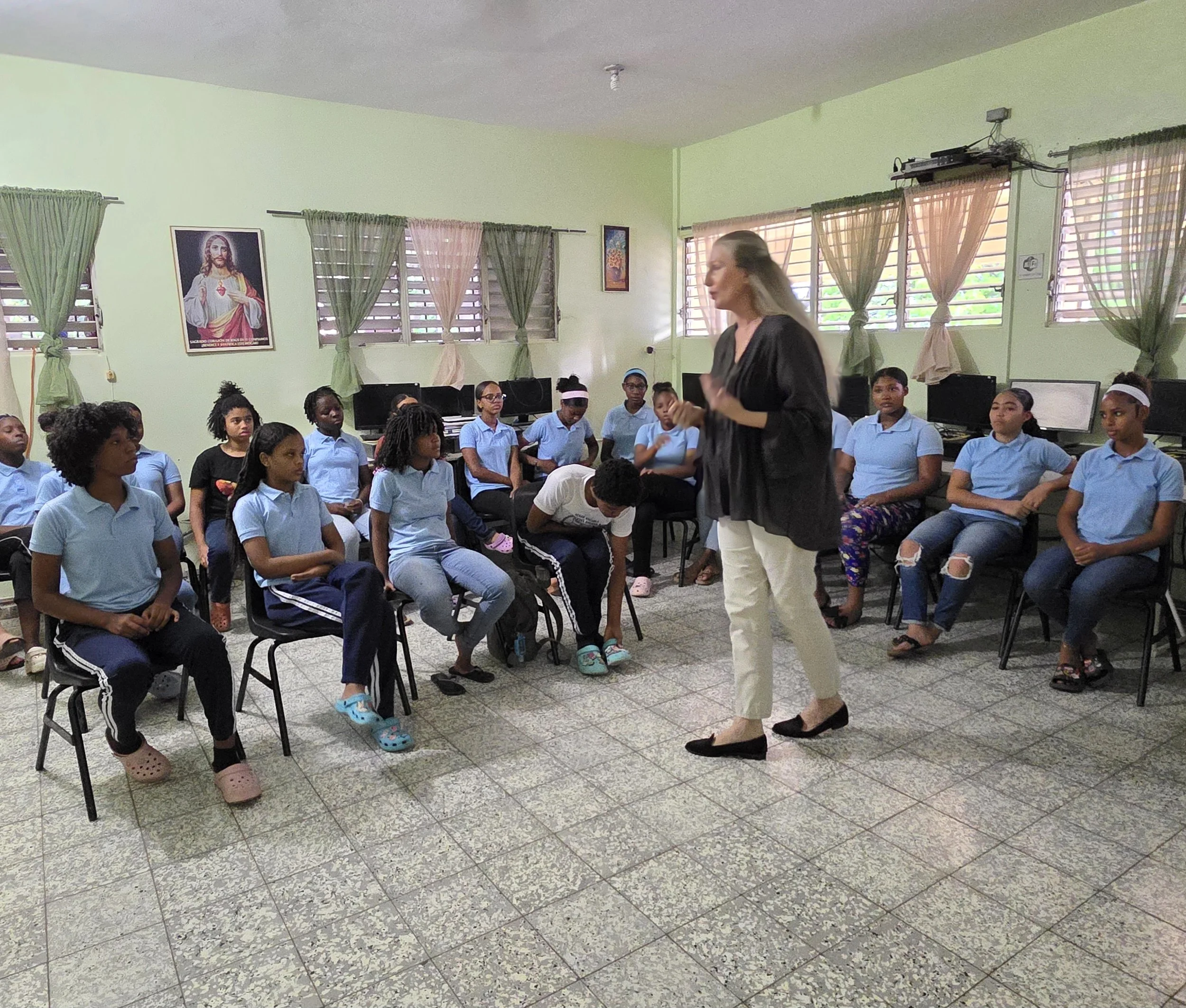 A classroom with a female teacher standing in front of a group of students who are sitting on chairs. The students are mostly girls dressed in blue uniforms. Some students are paying attention while others are looking away or adjusting their clothes.