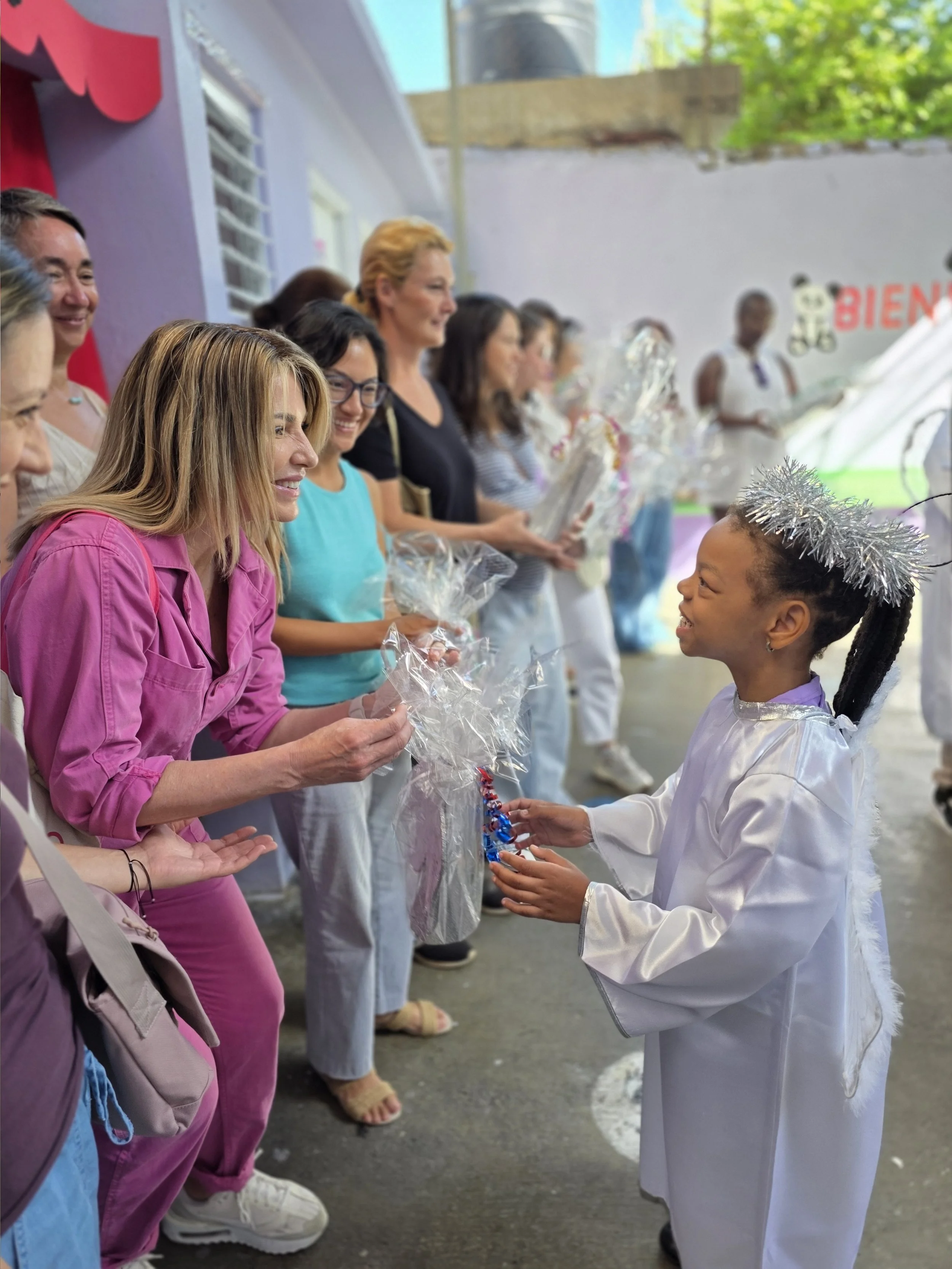 Young girl dressed as an angel receiving a gift from a woman during a celebration, with a line of people holding gifts in the background.