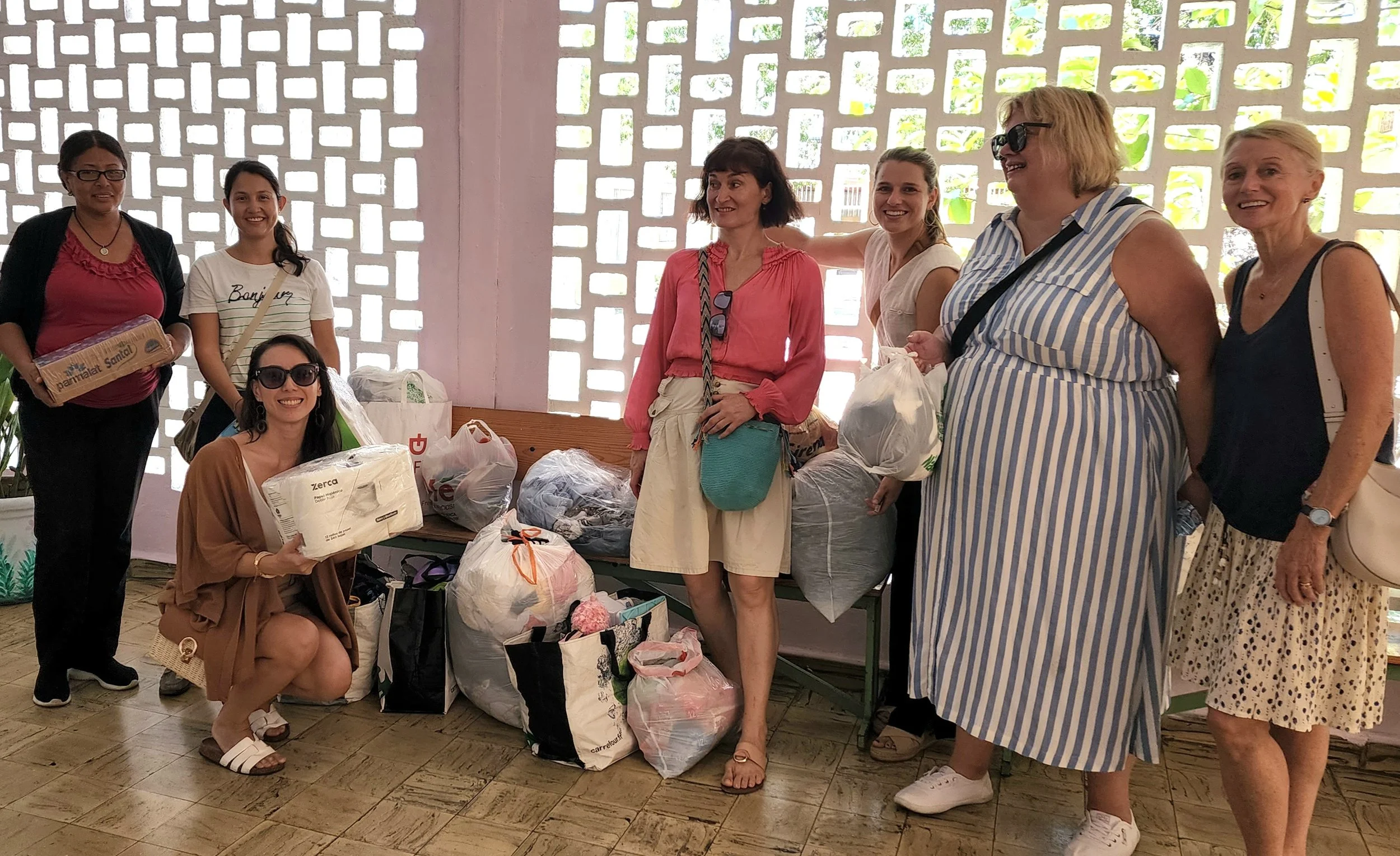 Group of women standing and squatting indoors with bags and packages of supplies, smiling and posing for the photo.
