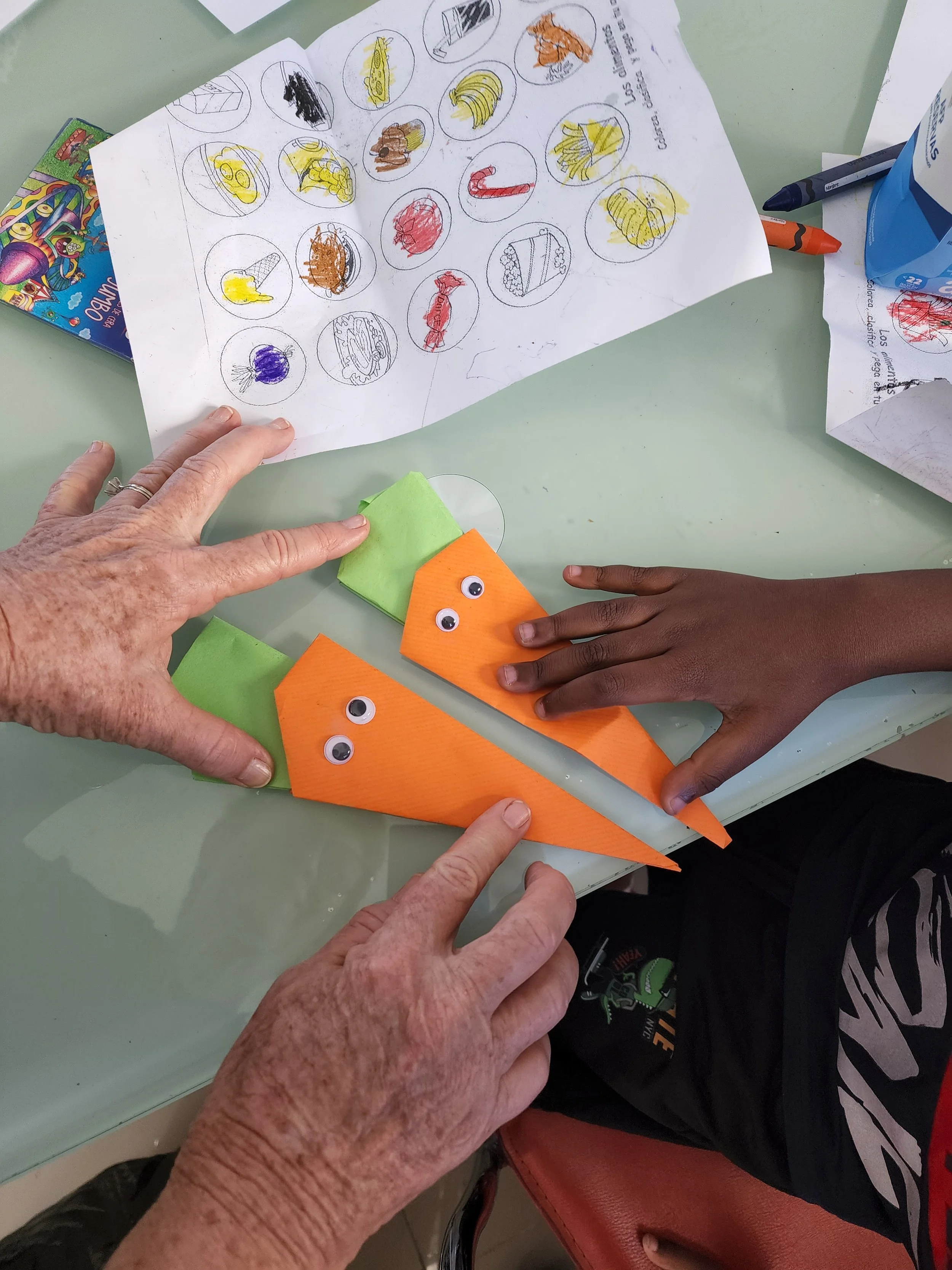 Two people making paper carrots with orange paper, green paper tops, and googly eyes. One person is an older woman with light skin, and the other is a young person with dark skin. They are at a table with coloring pages and markers.