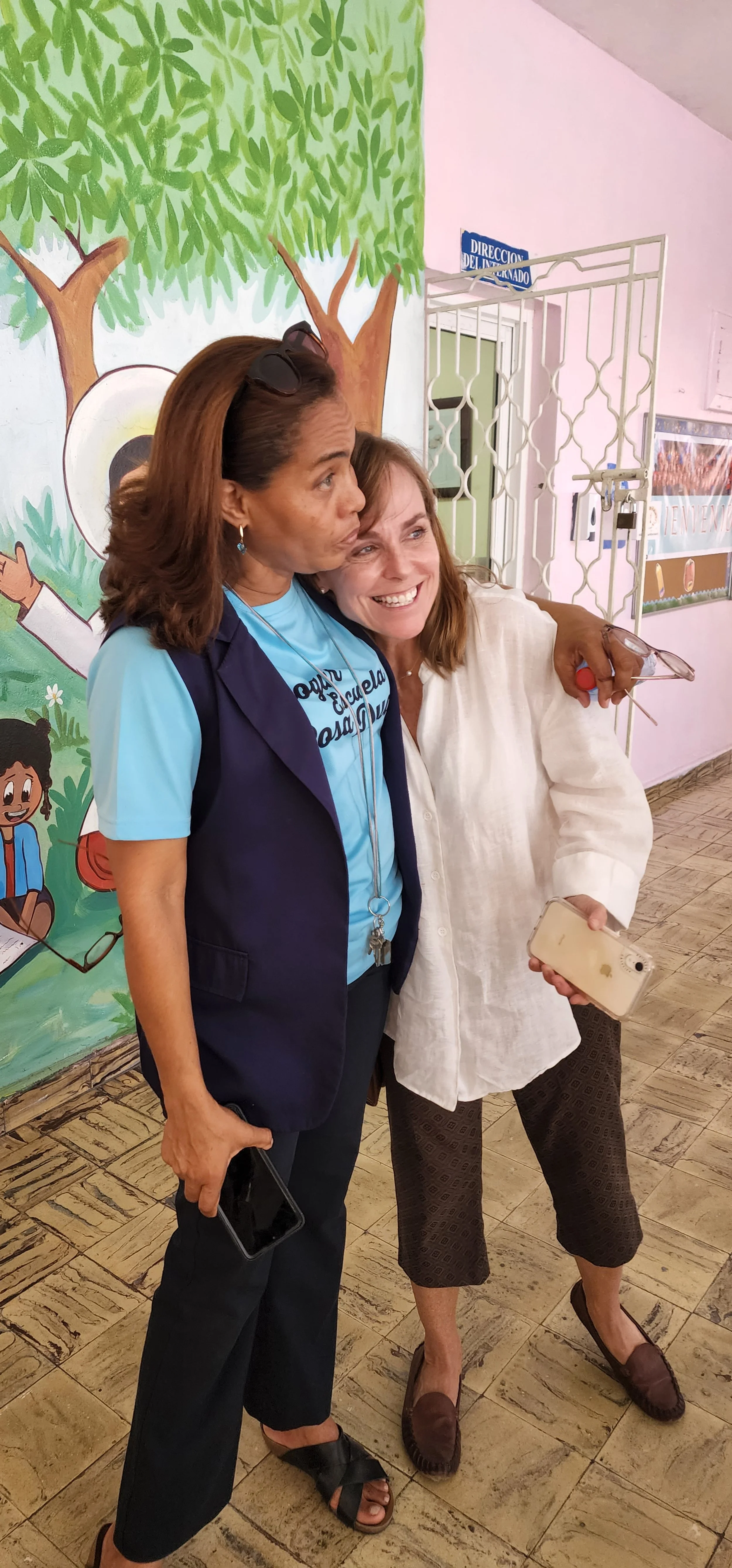 Two women hugging and smiling, standing in front of a colorful mural with trees and characters, inside a building.