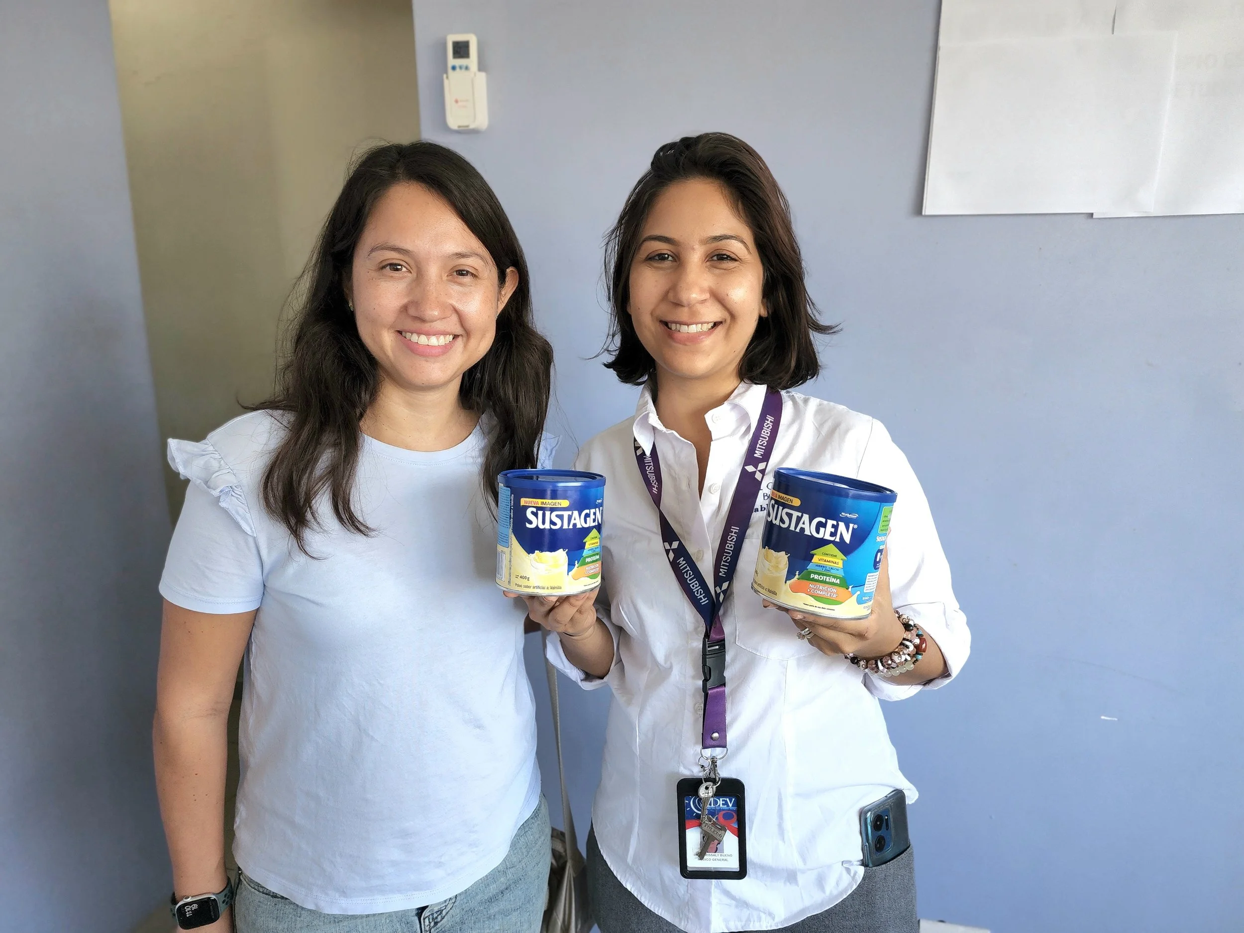 Two women standing together, smiling, each holding a can of Sustagen nutritional supplement. One woman wears a white shirt with ruffles on the shoulders, and the other wears a white shirt with a Mitsubishi lanyard and ID badge. They are in a room wit
