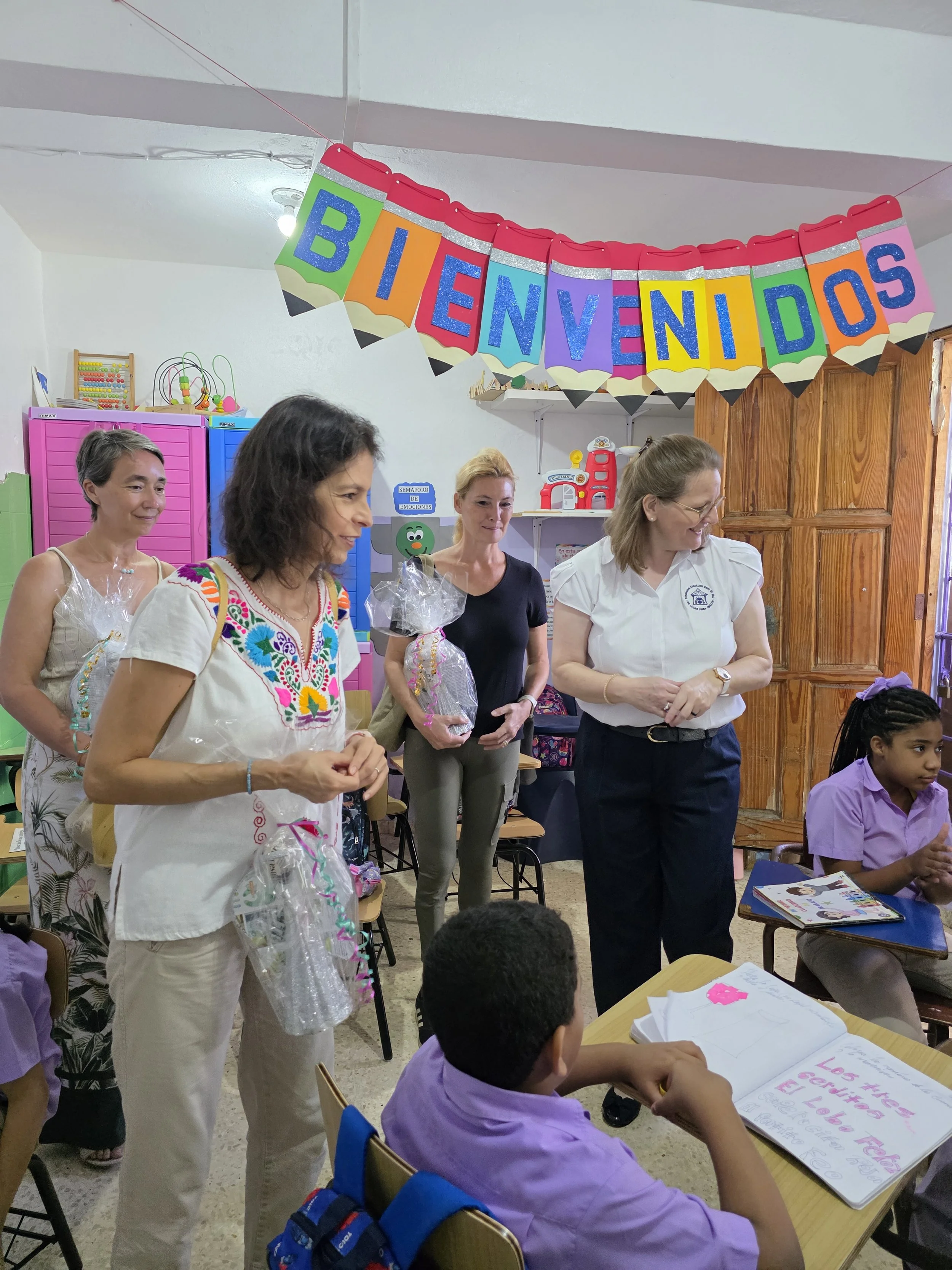 Women and children in a classroom decorated with a colorful 'Bienvenidos' banner, engaging in an activity.