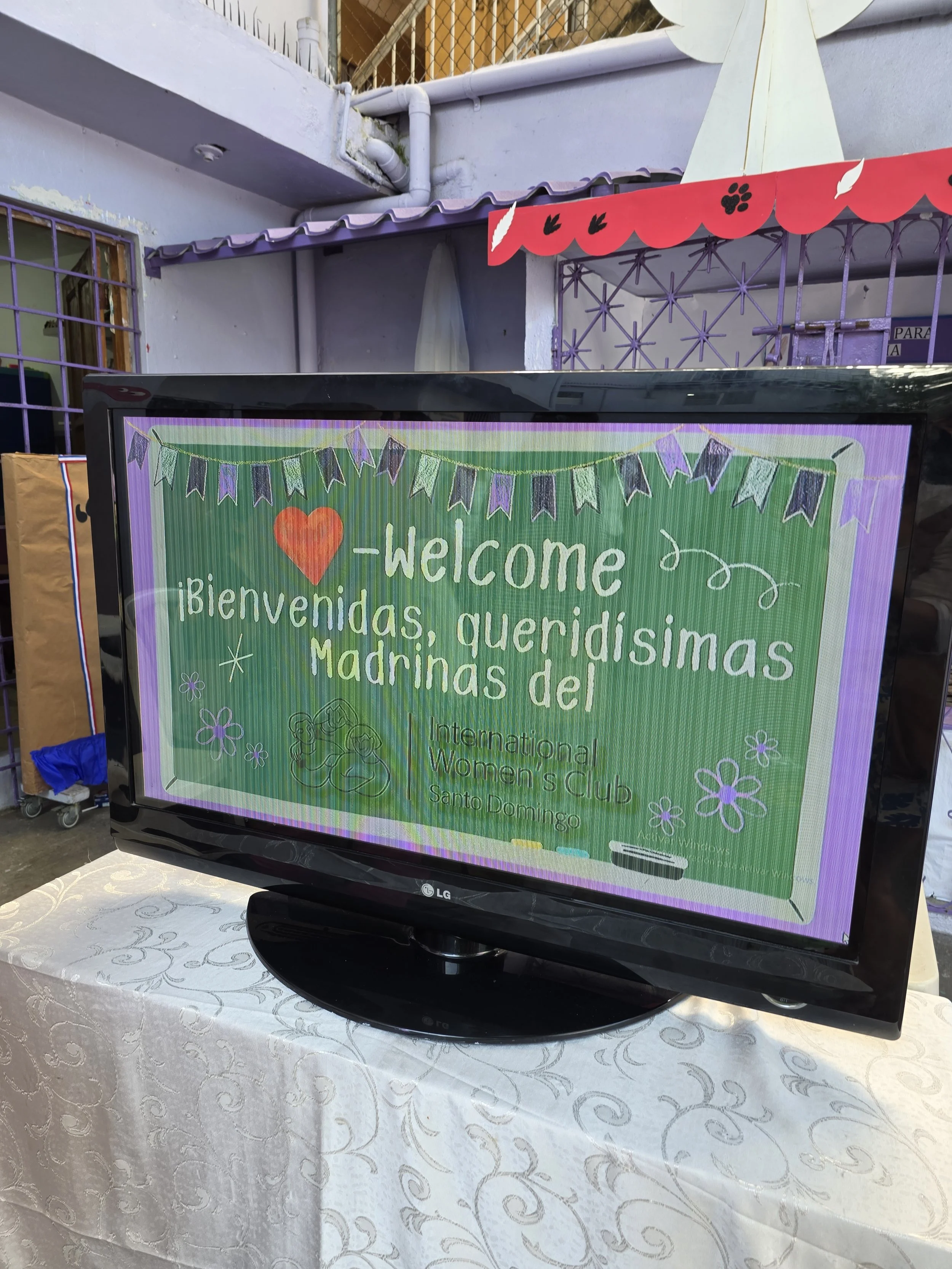 Television screen displaying a welcome message for the International Women's Club Santo Domingo in English and Spanish, decorated with flowers and banners.