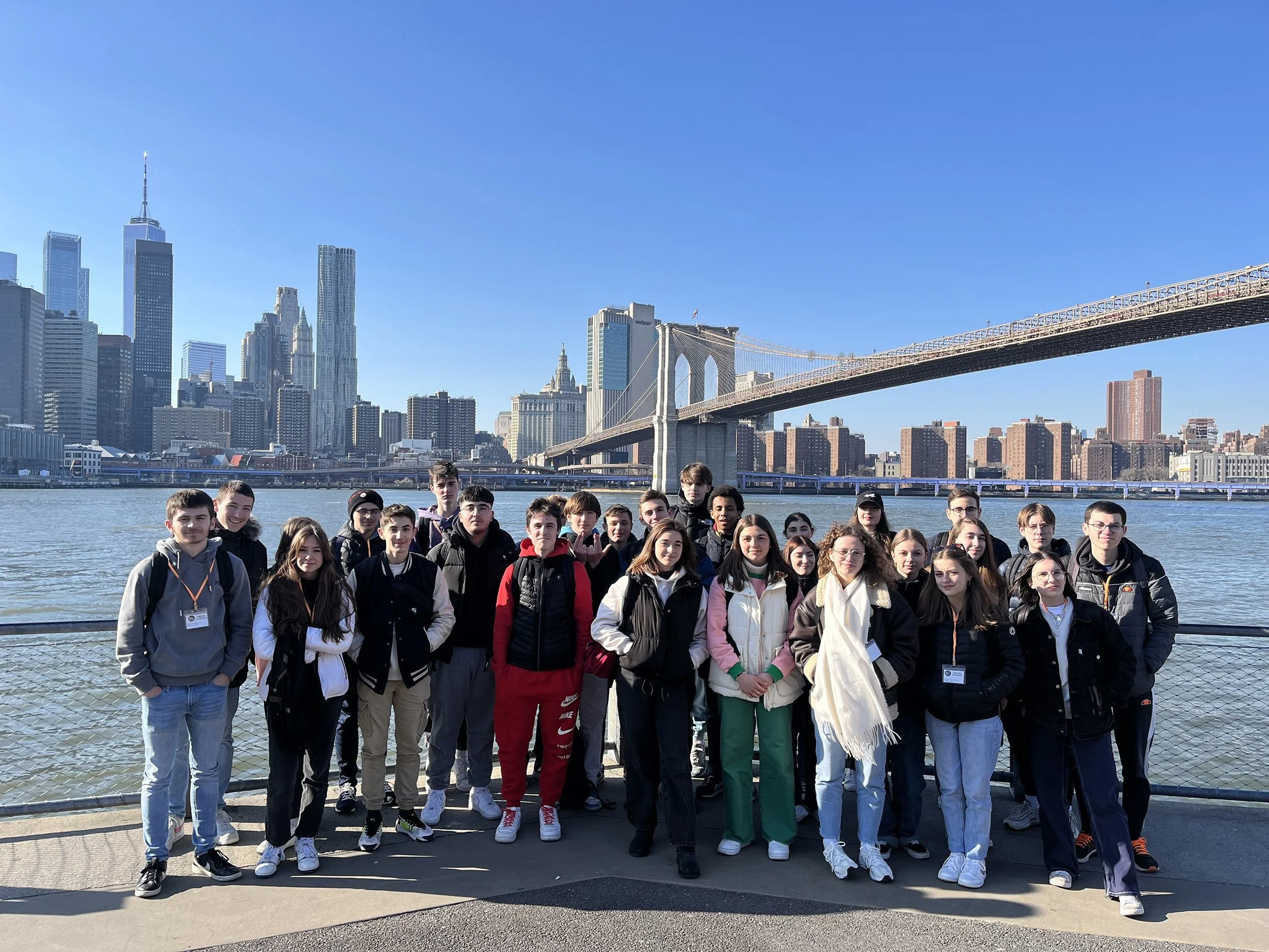 Group of young people standing by the water with the Brooklyn Bridge and Manhattan skyline in the background on a clear day.