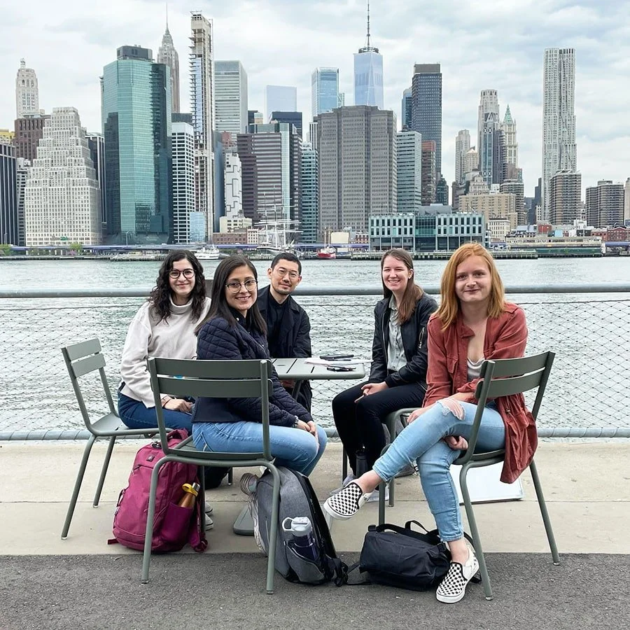 Five young adults sitting at a table outdoors with a city skyline in the background, near a body of water. They are smiling and appear to be enjoying a casual gathering.