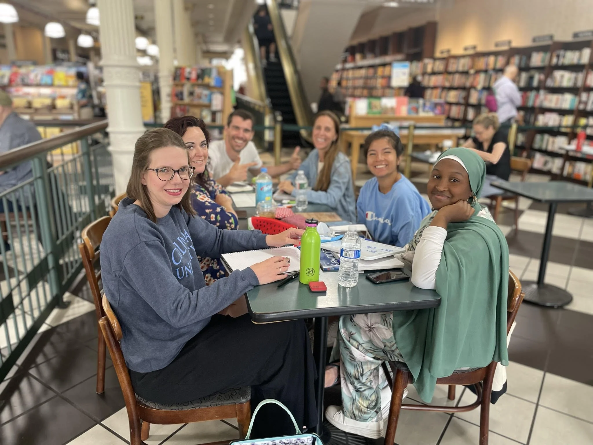 Six women sitting at a table in a bookstore or library, smiling and talking, with bookshelves in the background.