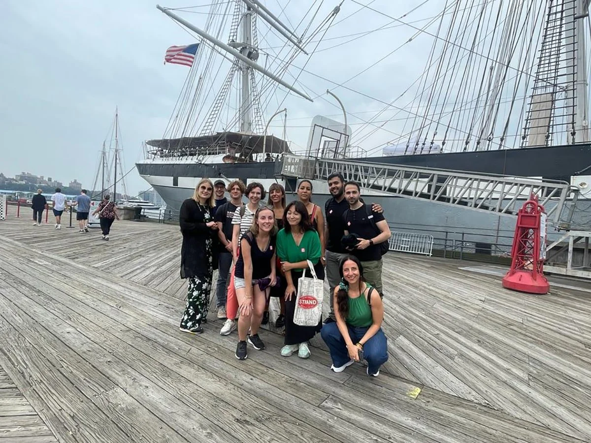 Group of people posing in front of a large sailing ship at a waterfront dock.