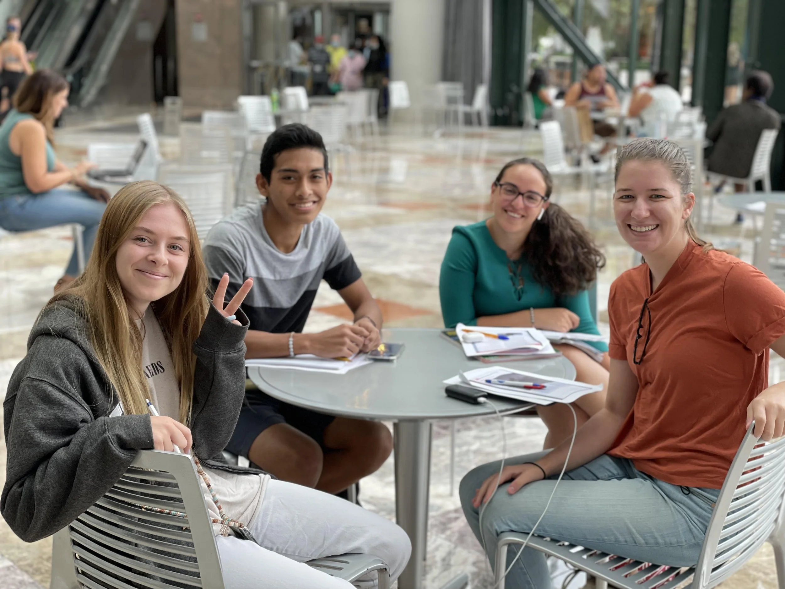 Four young women seated around a table at a cafe, smiling and posing for the camera. They have notebooks and pens in front of them, indicating they are studying or working together.