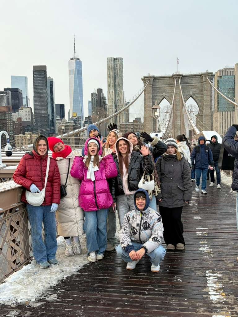 Two young girls wearing helmets stand with their bikes on a city street, covering their mouths in surprise or laughter, with other cyclists and pedestrians in the background.