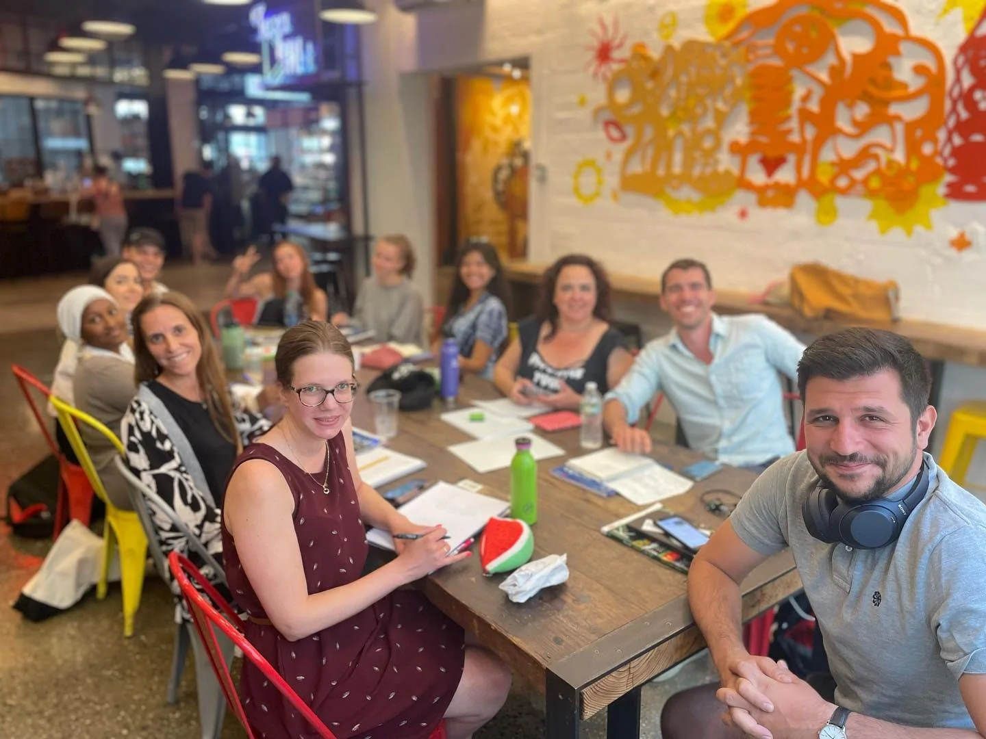 A group of people sitting around a large wooden table in a restaurant or cafe, smiling for the camera, with a colorful abstract mural on the wall behind them.