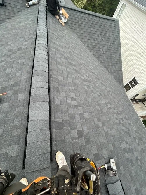 View of a sloped roof with asphalt shingles, showing workers installing or repairing the shingles near the ridge line.