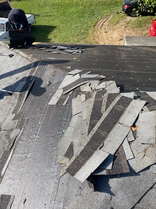 Broken asphalt shingles scattered on a roof with a person working nearby and a green lawn in the background.