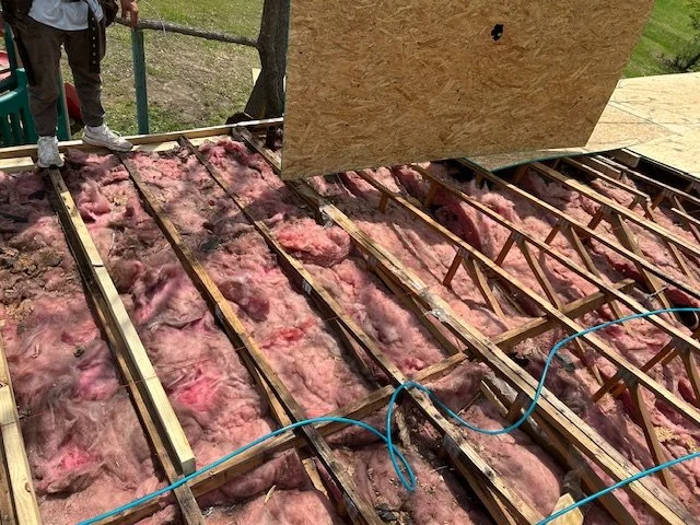 Construction site with pink insulation and wooden floor joists, with workers and tools visible.