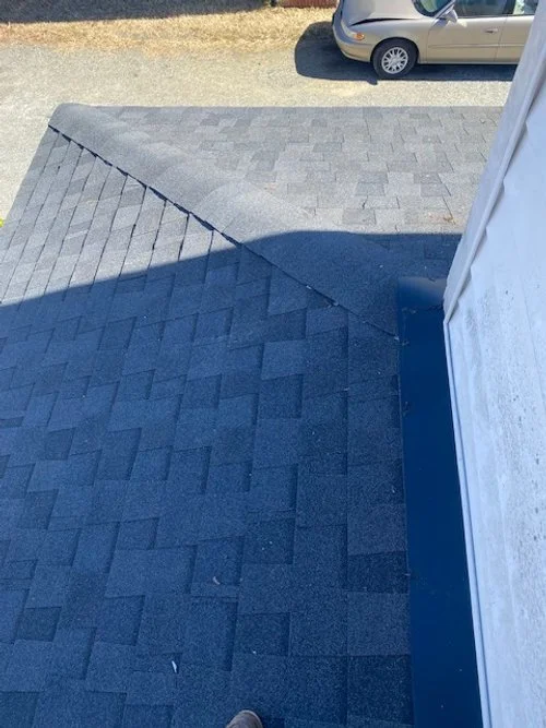 View of a sloped roof with asphalt shingles and a shadow cast over part of the roof, with a residential street and parked car visible in the background.