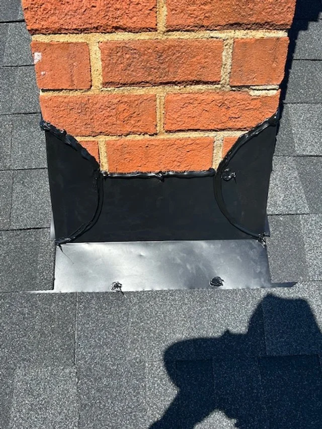 A brick chimney with a metal flashing and black waterproof sealant on a shingled roof.