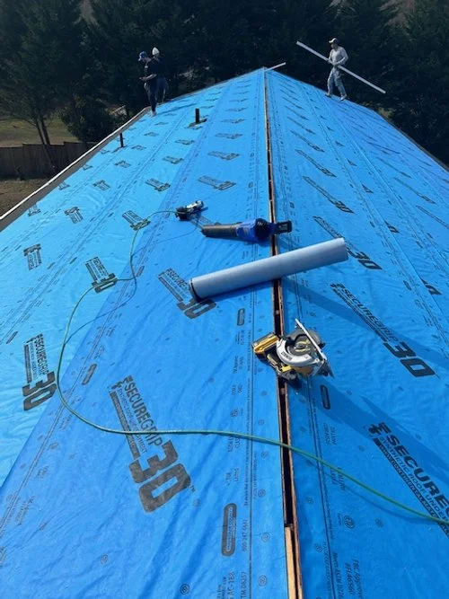Roof under construction with blue insulation, tools, and two workers installing the roof in the background.