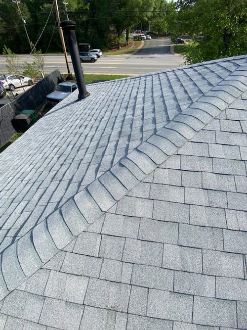 Upward view of a gray shingled roof with a chimney and antenna, overlooking a street with parked cars and trees.