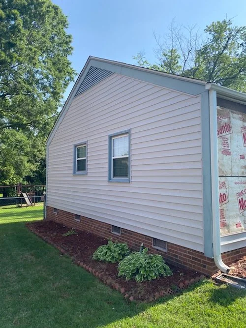 Side view of a house with beige vinyl siding, two small windows, and a brick foundation, with a small garden bed along the base and two green plants, under a clear blue sky.