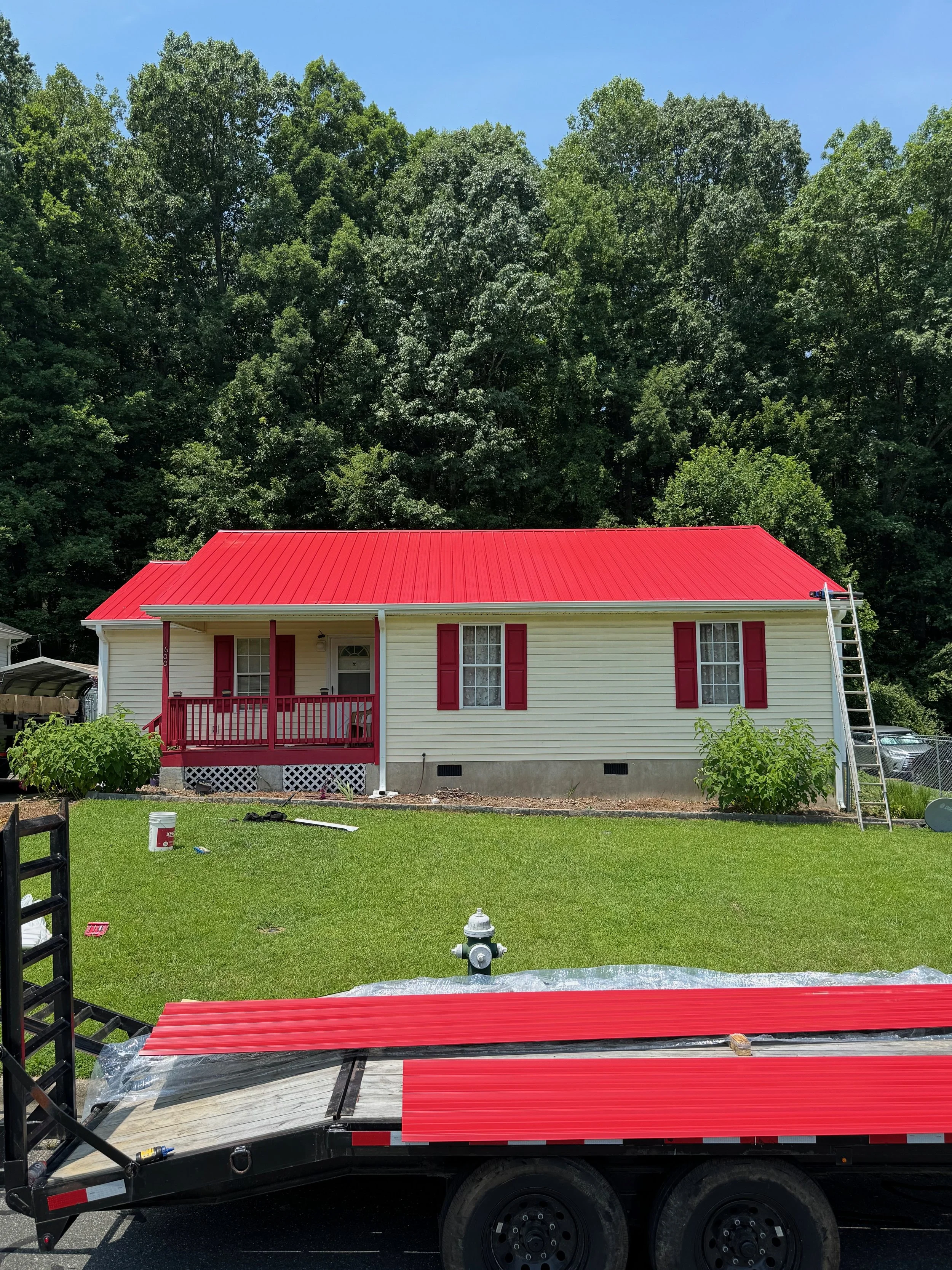 A house with a yellow exterior and red roof, surrounded by green trees, with a trailer on the driveway containing red roofing sheets and tools.