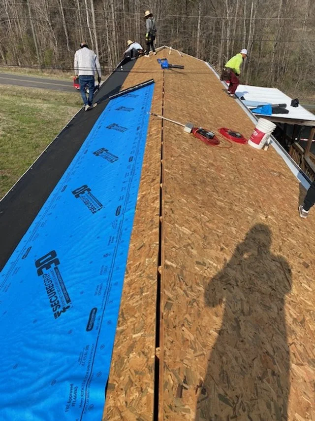 Workers installing roofing on a house, with plywood sheathing and blue weatherproofing underlayment visible.