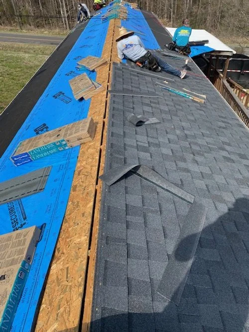 Workers installing asphalt shingles on a roof with roofing materials and tools visible.