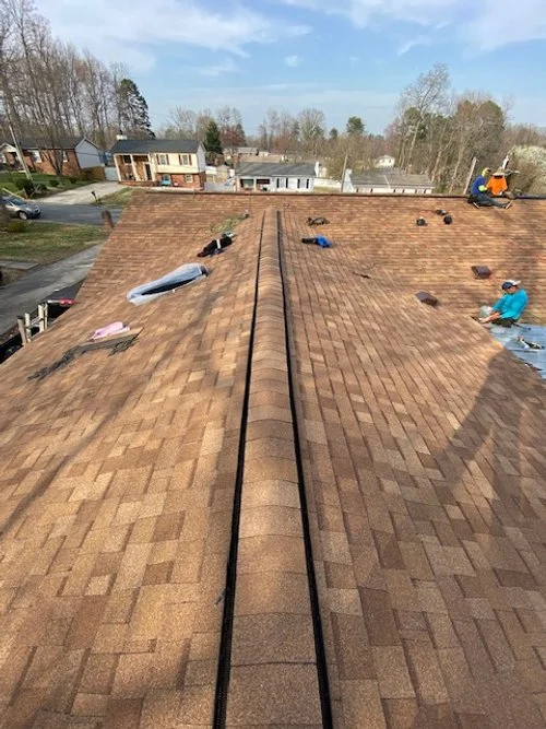 View from the peak of a house roof with shingles, showing roofers working and equipment on the shingles.