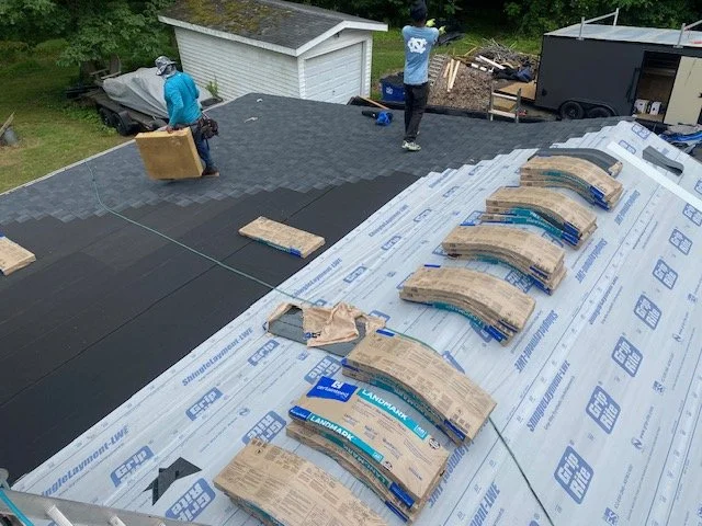 Two workers installing black roofing shingles on a house roof, with shingles and underlayment materials laid out nearby.
