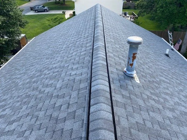 View of a gray shingled house roof with a vent pipe showing rust and peeling paint, and a small black cable or wire running along the peak.