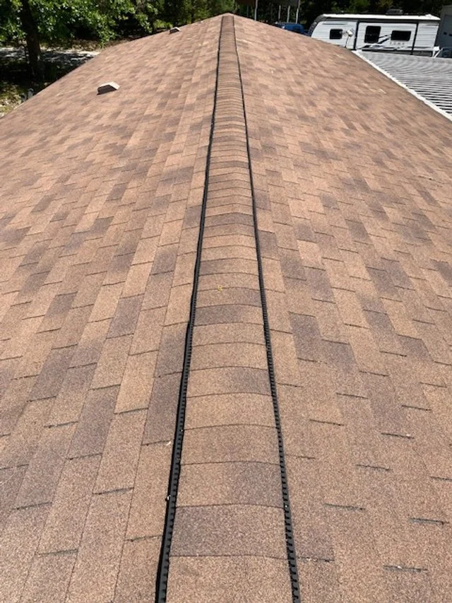Close-up of a house roof with brown asphalt shingles and a vent pipe, showing the central ridge and ridge vent.