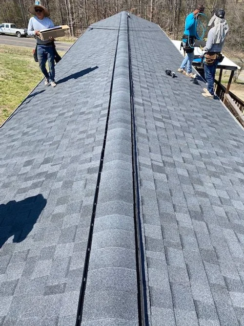 Roof with gray asphalt shingles being inspected and worked on by three people wearing helmets and safety harnesses.