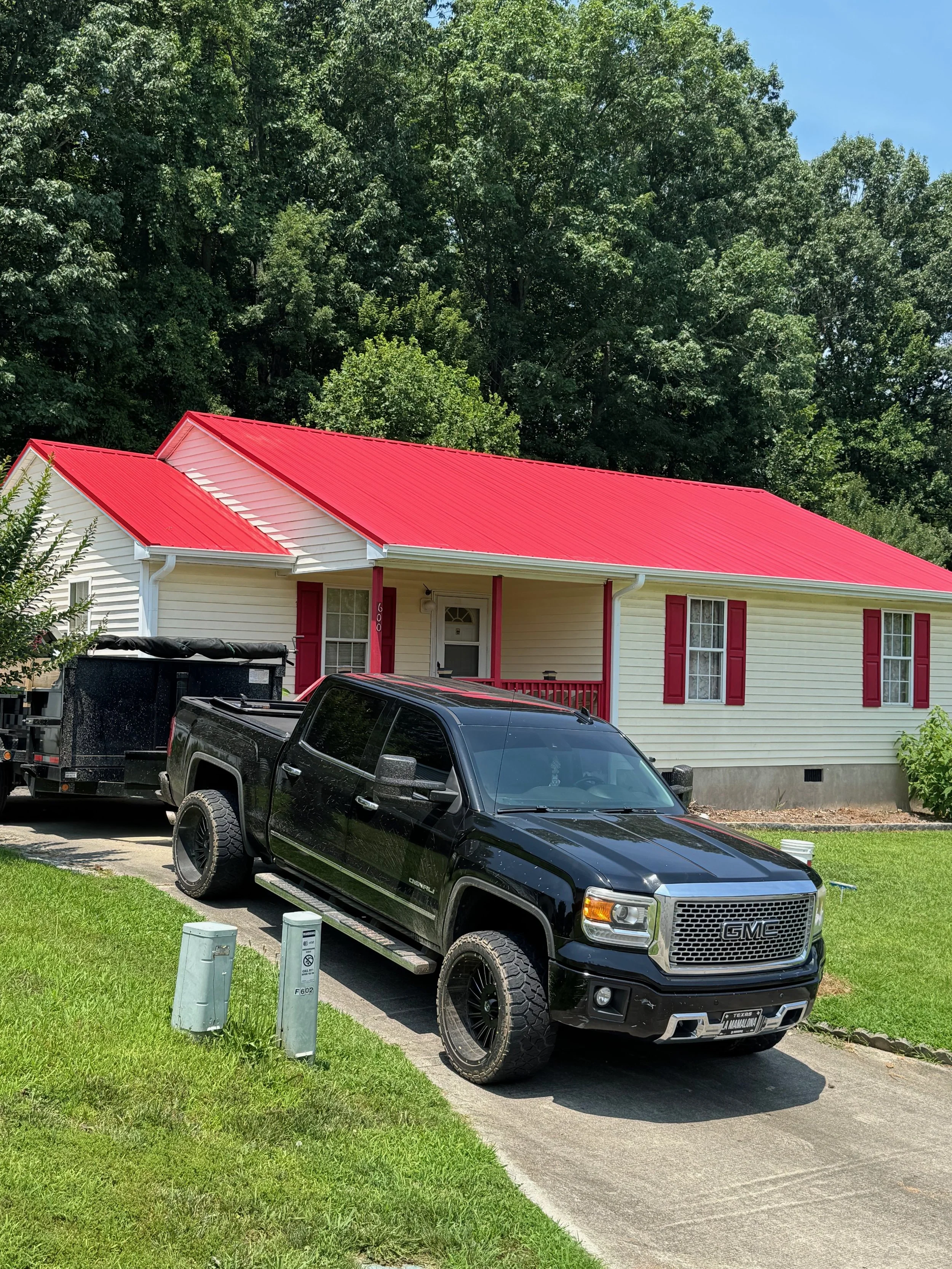 A black GMC truck parked on a driveway in front of a white house with red shutters and a red metal roof, surrounded by green lawn and trees.