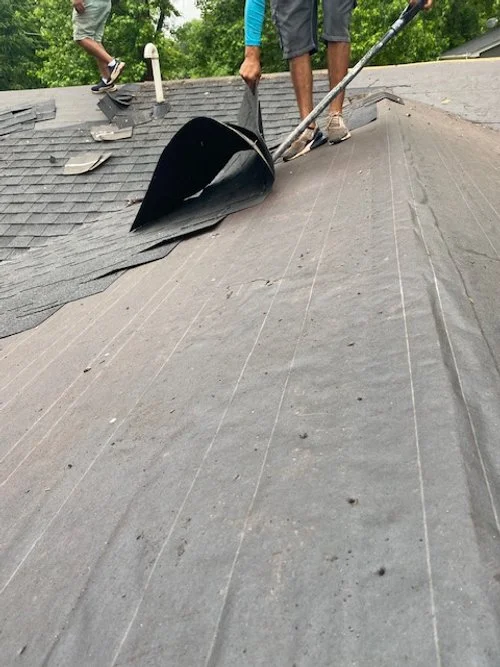 Two workers installing or repairing roofing shingles on a sloped roof using a large pry bar or tool.