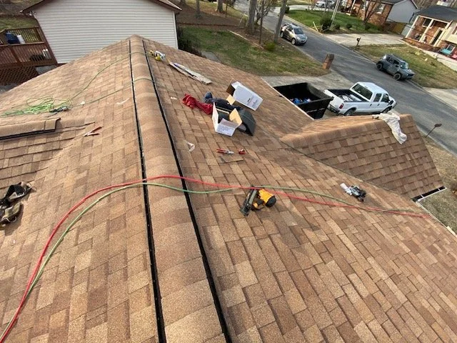 Peak view of a rooftop under construction or repair, with tools, boxes, and equipment scattered on the shingles, and surrounding neighborhood streets visible in the background.