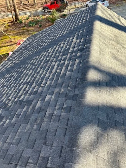 View of a gray shingle roof from above with shadows cast across the shingles, and a yard with trees, bushes, and cars in the background.