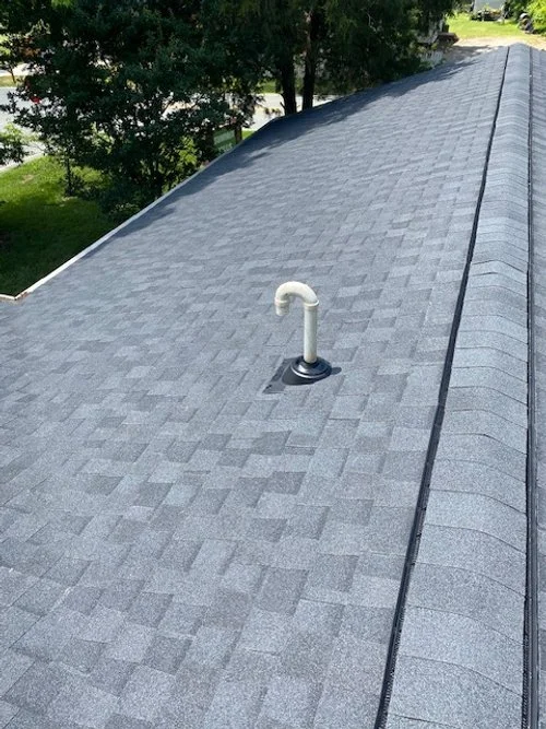 View of a gray shingled rooftop with a vent pipe and lush green trees in the background.