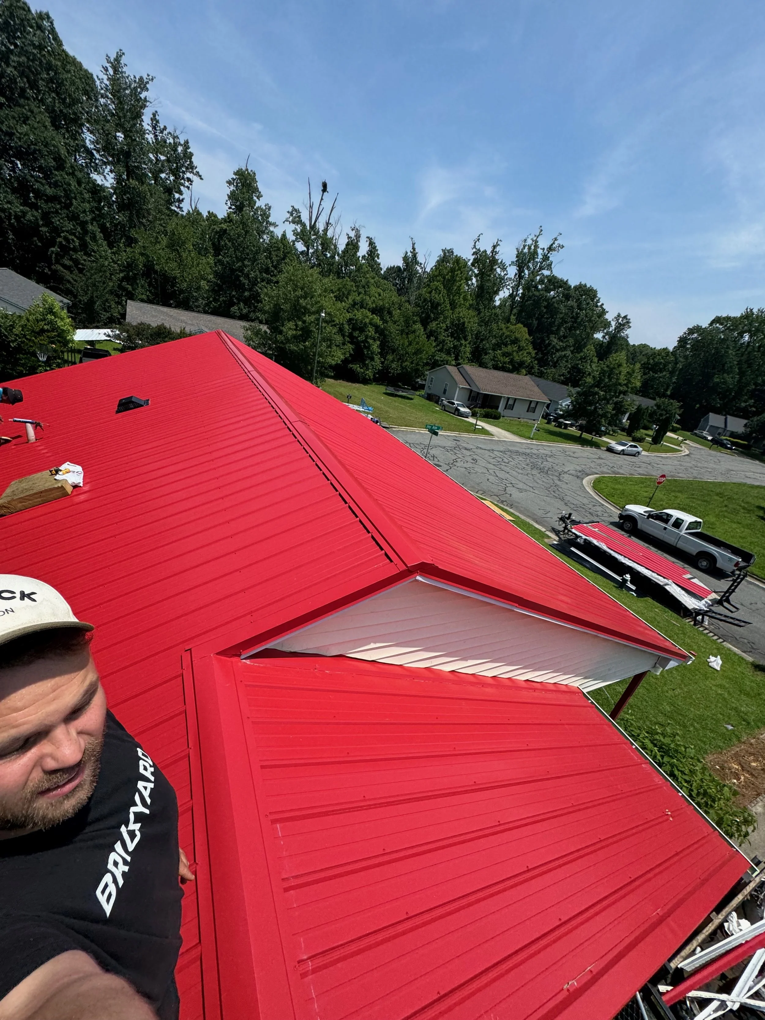 A person standing on a ladder or roof taking a selfie, with a bright red metal roof and a suburban neighborhood in the background.