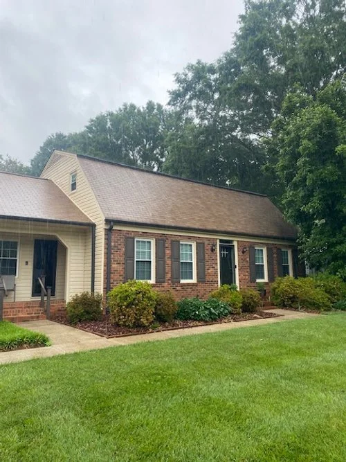 Front view of a brick and siding house with green lawn and bushes, overcast sky, and trees in the background.