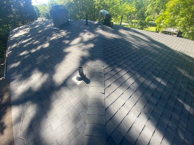 View of a shingled roof with shadows from trees, a chimney, and vent pipes visible.