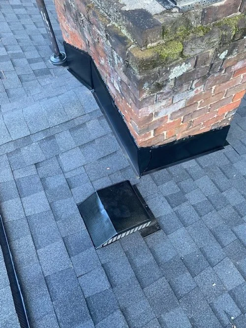 Close-up of a brick chimney with a black flashing at its base on a shingled roof, with a small black metal vent or cover on the shingles nearby.