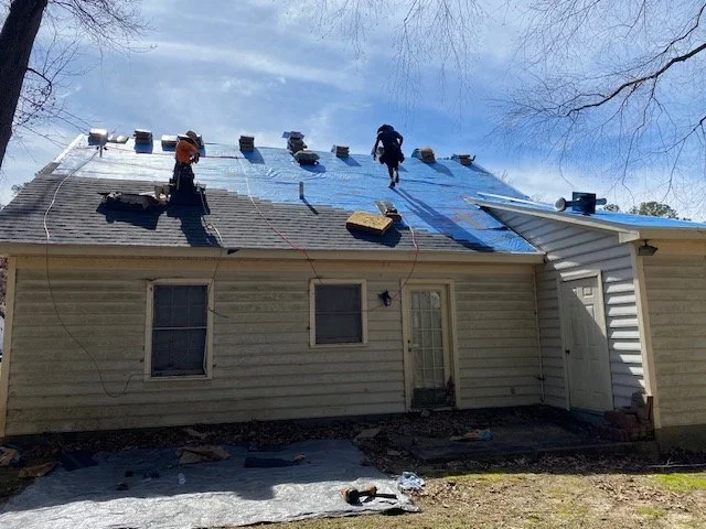 Two workers installing a blue metal roof on a house under a partly cloudy sky.